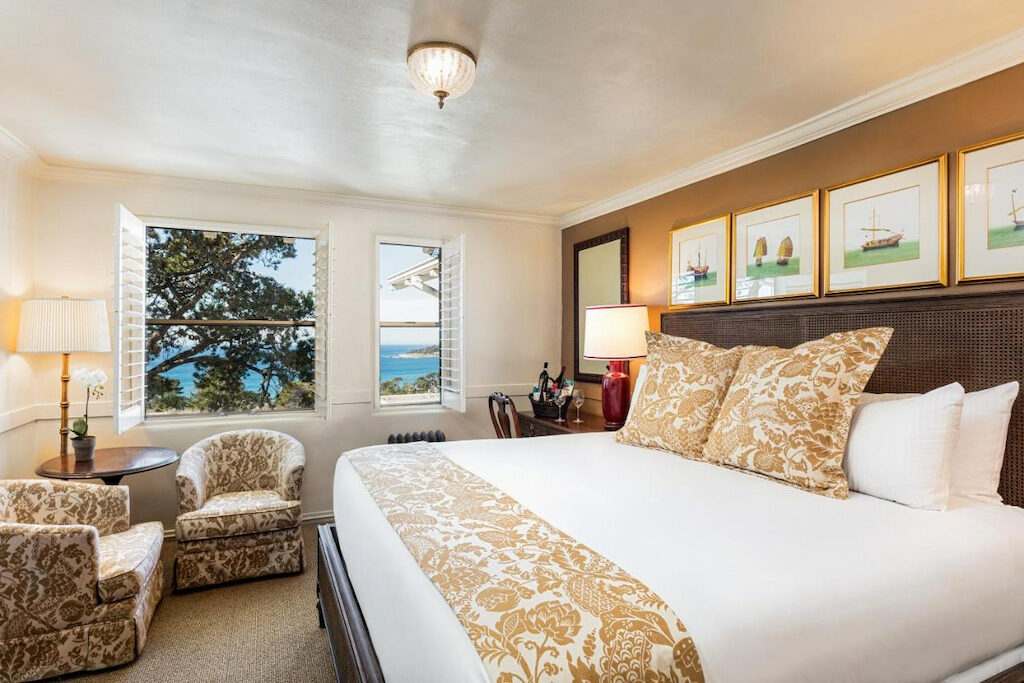 A carpeted room in one of the boutique hotels in Carmel by the sea with a white queen bed, chairs, tables, lamps and open windows with a view of the sea 
