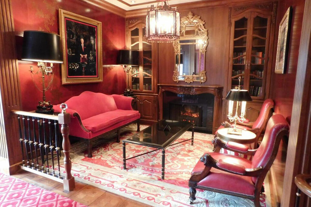 A carpeted, antique-style room with red couch and chairs, rectangular and circle tables, close shelves, fireplace, mirror, and framed portrait