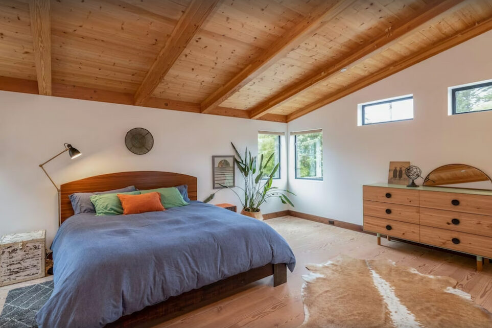 A room with wooden ceiling and a king size bed with blue sheets near a wooden drawer and a couple of small windows in one of the hotels in Half Moon Bay.
