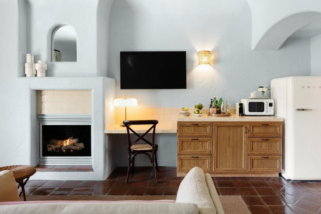 A white room with brown tiled floor, fireplace, couch, lamps, chair, flat-screen television, and a kitchenette with a white refrigerator and a counter with a microwave and supplies