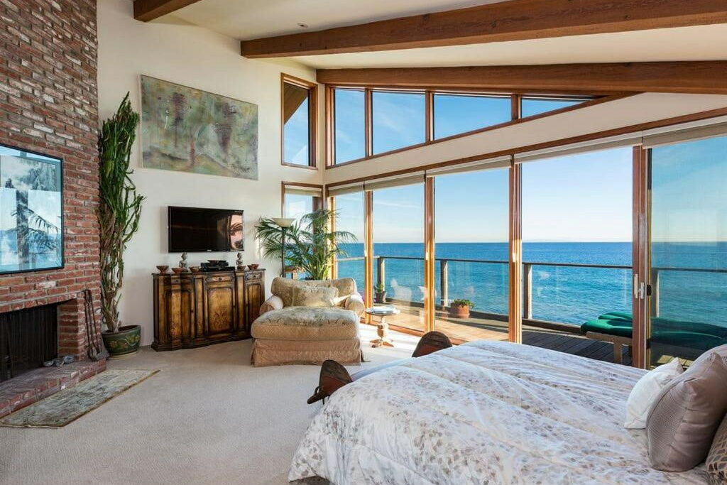 A room with ceiling-to-floor windows with a cozy bed, fireplace, couch, flat screen TV and view of the beach.