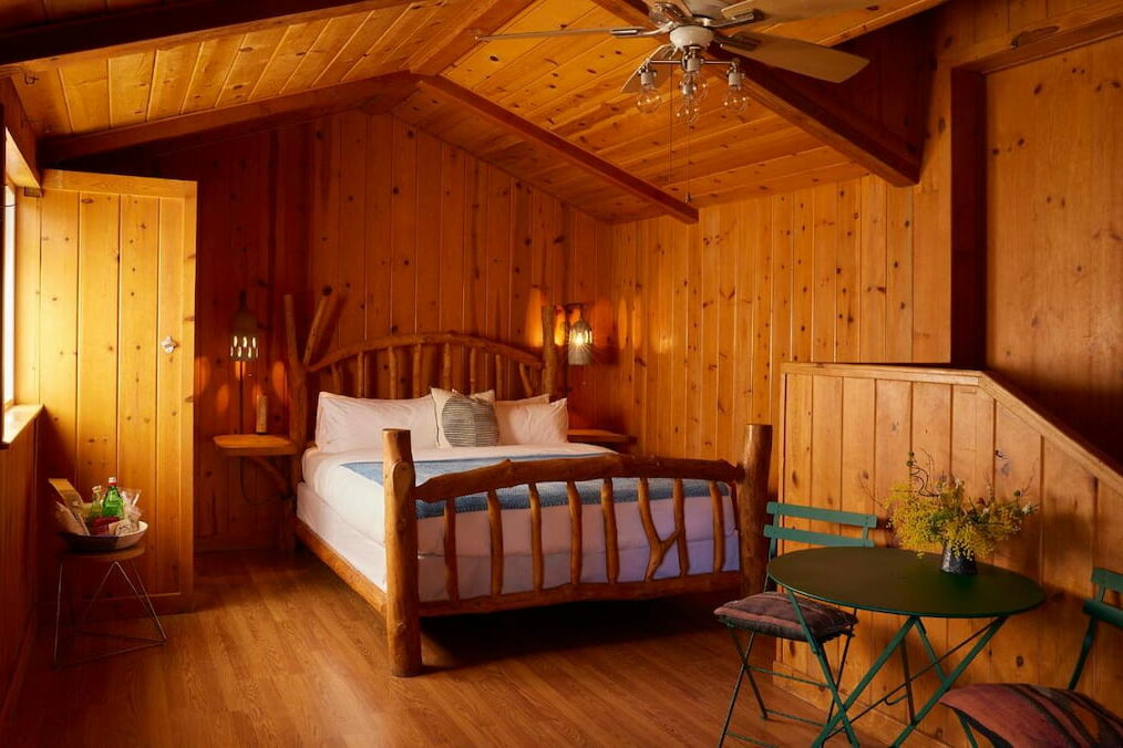 An all wood room with a cozy bed, small table and chairs and a ceiling fan.