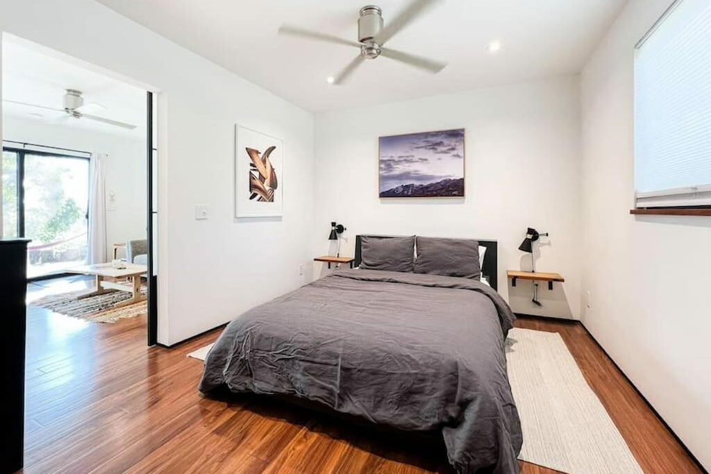 A spacious bedroom with. abed with gray sheets below a ceiling fan.