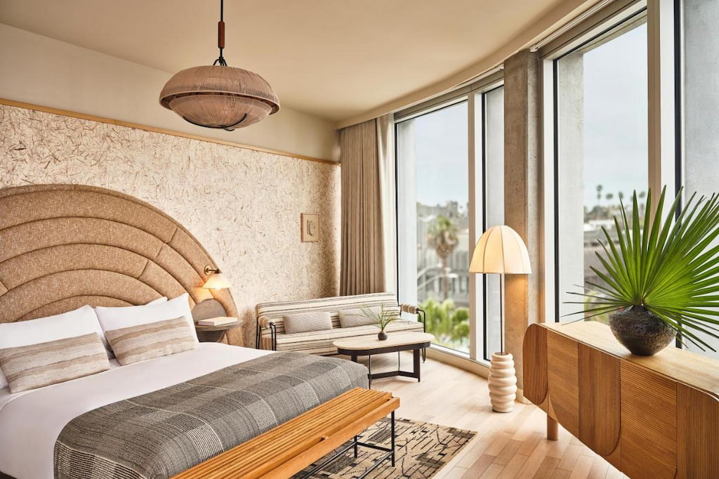 One of the luxury hotels in Venice Beach CA with a cozy bed with huge headboard, a lamp, small sofa, drawer and a ceiling-to-floor windows.