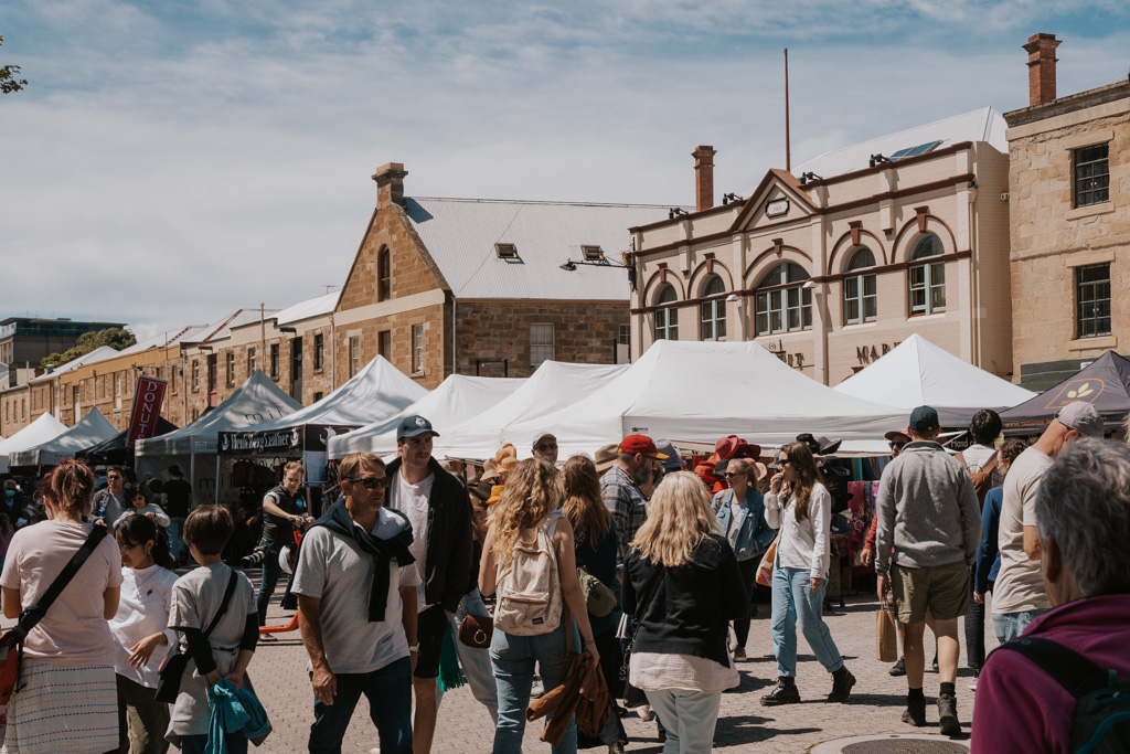 many people wandering around in a market with white topped stalls and heritage brick buildings in the background in Hobart's CBD Salamanca area