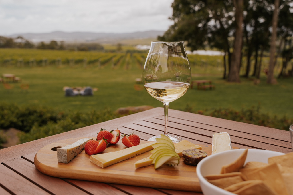 a wooden platter with small chunks of various cheese and strawberries and apple with a glass of white Tamar Valley wine and vineyards in the distance