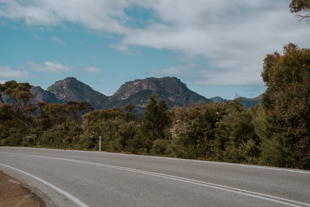 a paved road lies from right to left of the image with short trees running along side and rugged mountain peaks in the distance on a partly cloudy day in Freycinet National Park along a road trip east coast Tasmania