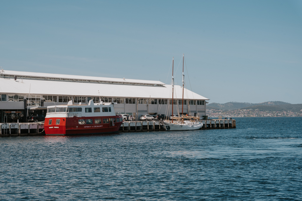 red and white passenger boat moored at a long white building on a wharf with water in the foreground in Hobart harbour