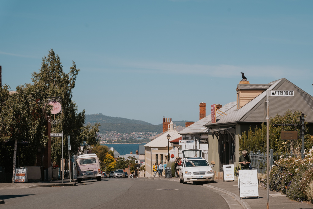 road with heritage buildings on each side and parked cars with ocean and mountains in the background on a blue sky day along a Hobart to Launceston road trip