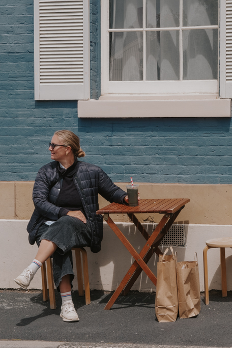 blonde woman in black clothing sits holding a drink at a wooden table with blue brick building as a background on a Tasmania east coast itinerary