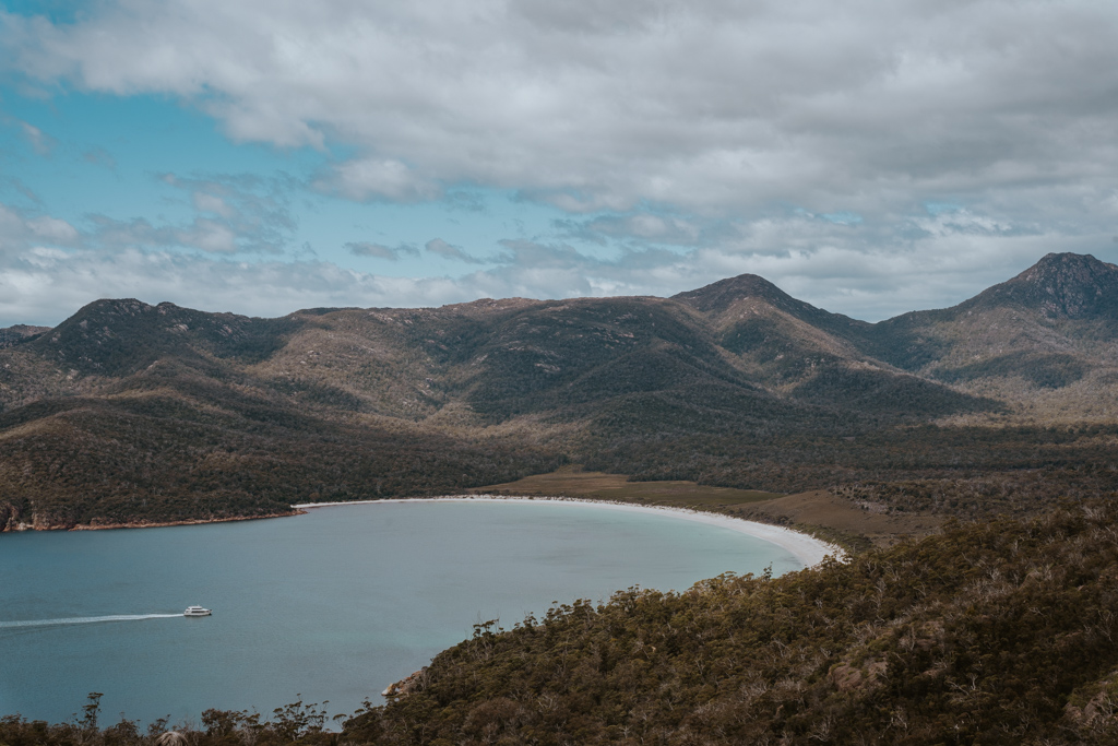 a small white boat cruises into a half moon shaped bay with bright blue water and undulating mountains in Wineglass Bay