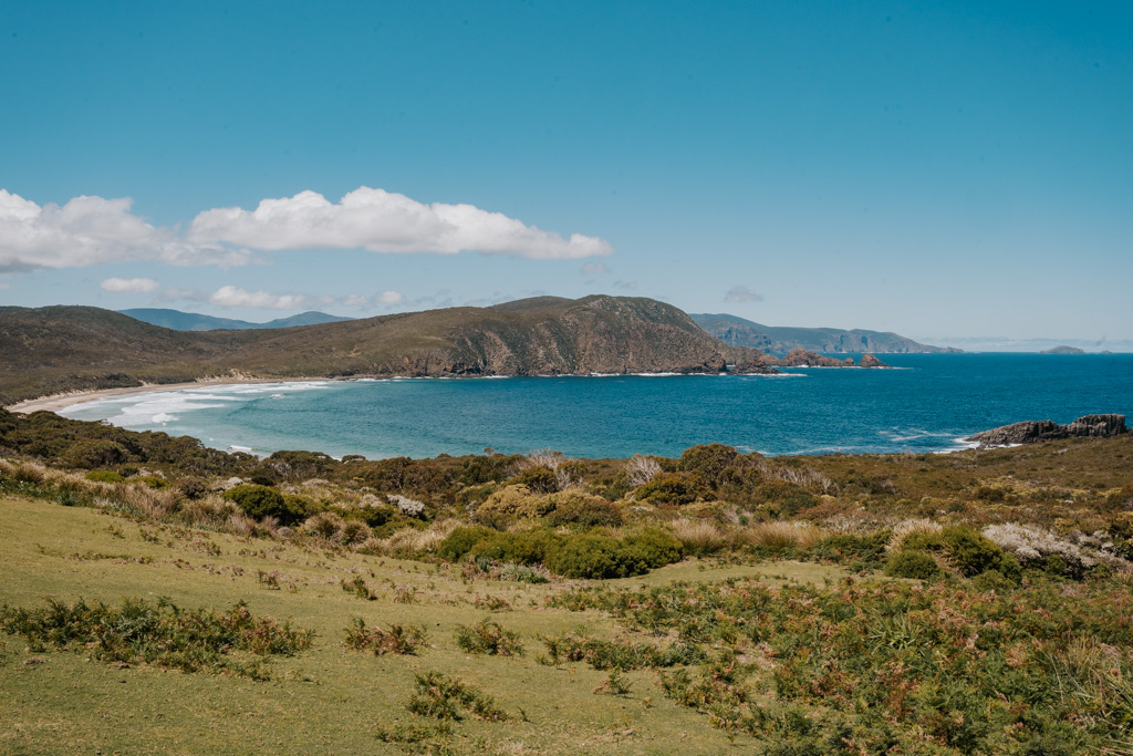 curved bay with bright blue water and white sand beach with low shrubbery and fields in the foreground in Bruny Island along a Launceston to Hobart road trip itinerary
