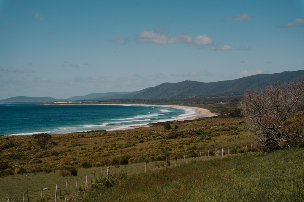 half moon shaped bay with white sand beach and green fields with mountains in the distance on a east coast Tasmania road trip itinerary
