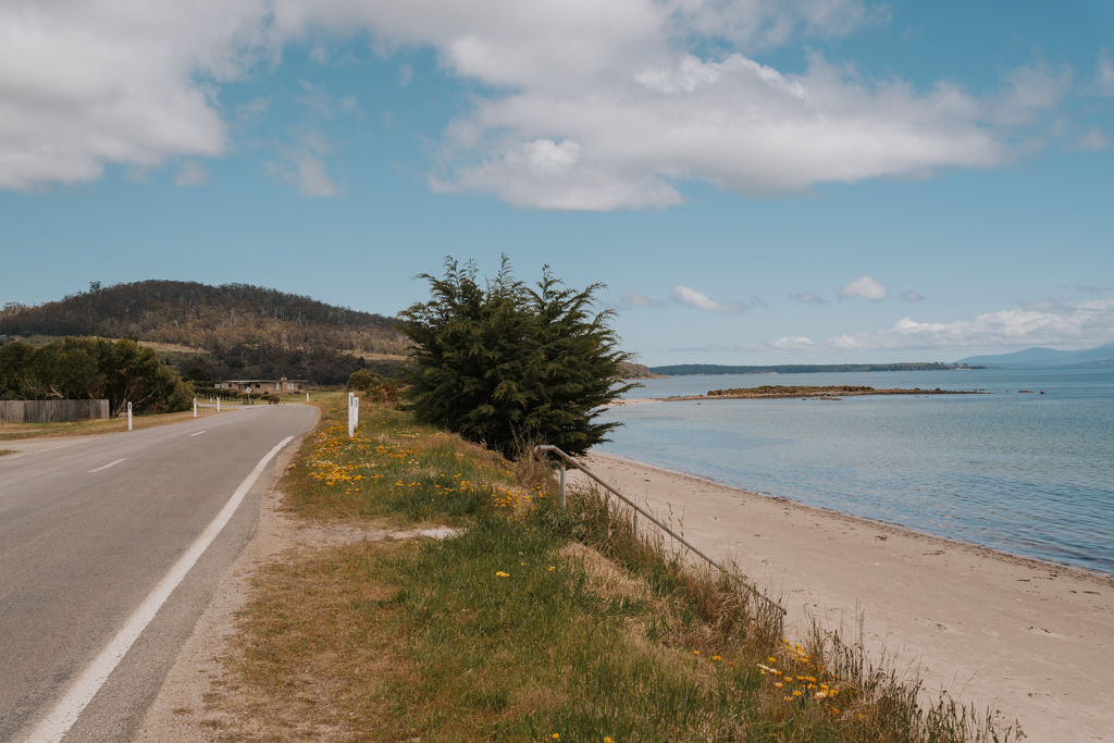 paved road meandering into the distance on the left with sandy beach and ocean to the right on Bruny Island a great destination to explore by car hire in Tasmania
