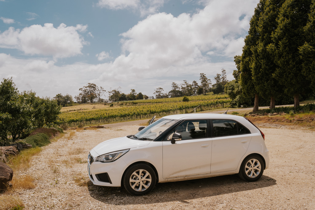 a white Tasmania rental car sits in a gravel parking lot in the foreground with green Tamar Valley vineyards beyond