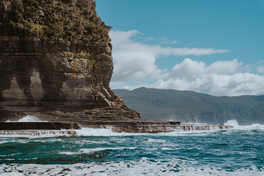 turbulent ocean water laps over tiered rocks on the shoreline with large cliffside on the left on a sunny day on a itinerary for Tasmania