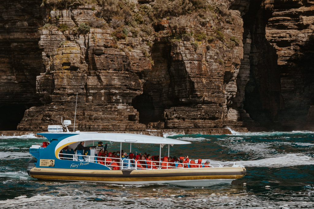 an blue adventure cruise boat with people wearing red jackets sits on the ocean in front of a dramatic rock cliff on their way to Tasman Island