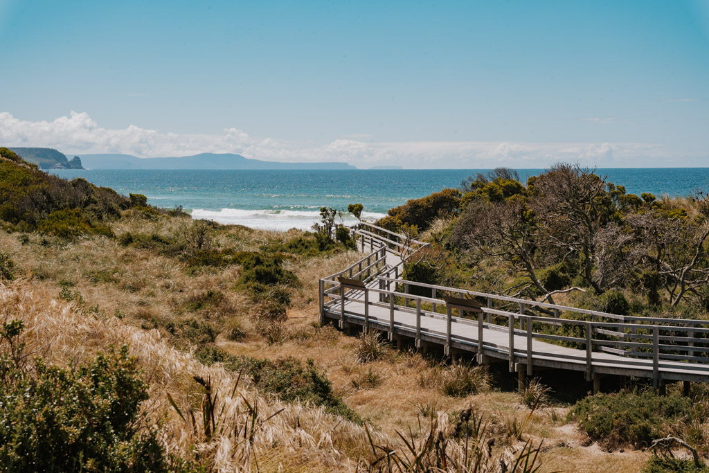 wooden boardwalk curves through a coastline green landscape toward a white sand beach with blue water