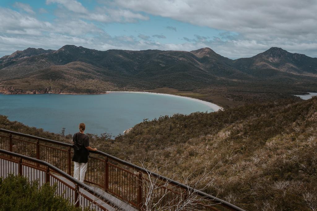a woman with a black shirt and white pants looks off in the distance from a boardwalk down to Wineglass Bay
