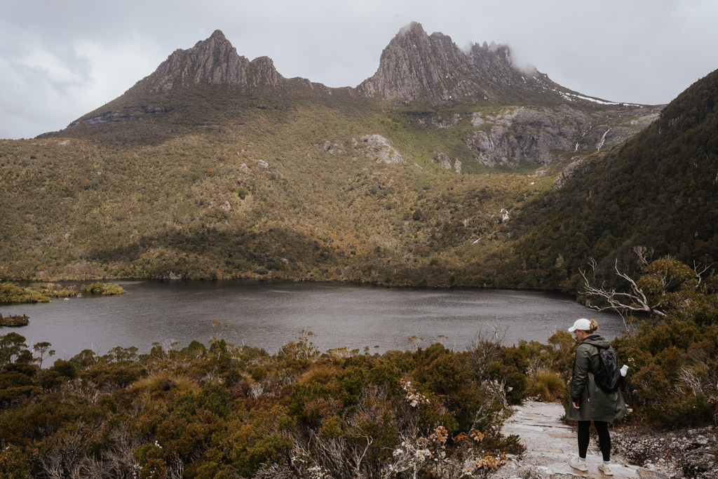 a girl with white hat and green rain jacket walks along Dove Lake with a looming Cradle Mountain peak in the background