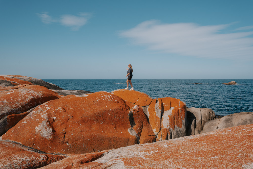 girl with blue shirt and black shorts walks across orange coloured boulders on the oceans edge in Bay of Fires