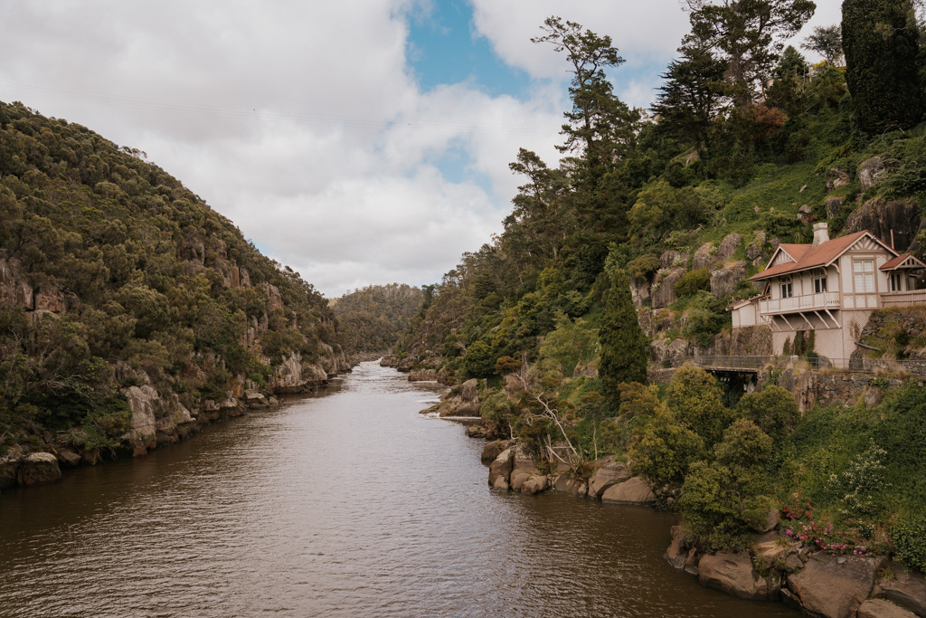 a white buildings with red roof overhangs the side of a canyon that has a river meandering off into the distance to Cataract Gorge