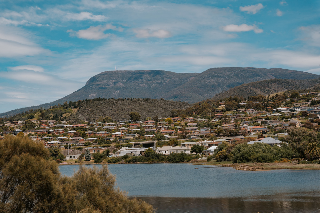 panoramic view overlooking an ocean inlet with a spattering of colourful houses on a green hillside with Cradle Mountain looming in the distance