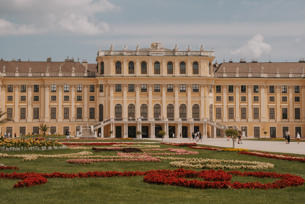 a three story classical palace building stands behind a manicured garden with low lying flowers in Vienna Austria one of the best places to visit in Europe for First time visitors looking for history