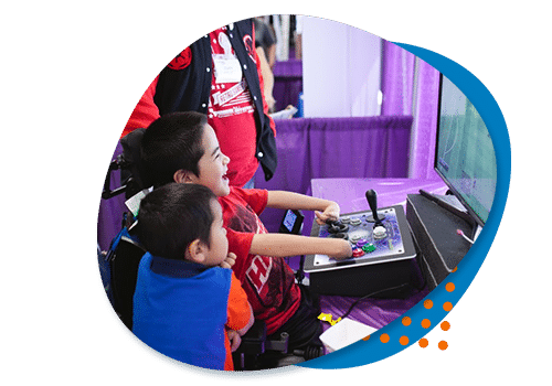 A royal blue roundish blob background small orange circles on top and on top of that a photo of a little boy laughing in a red t-shirt playing a video game using an adaptive controller while his little brother looks on