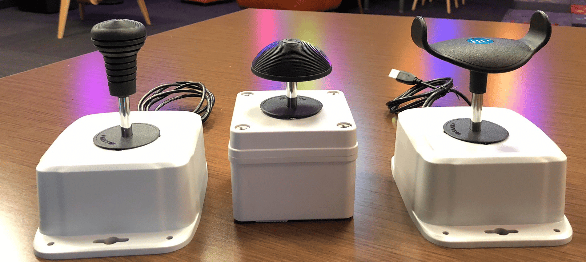 Three joysticks mounted to white boxes. The left one has a standard black joystick top, the middle one has a black, dome shaped top, and the right one has a black, u-shaped top. 