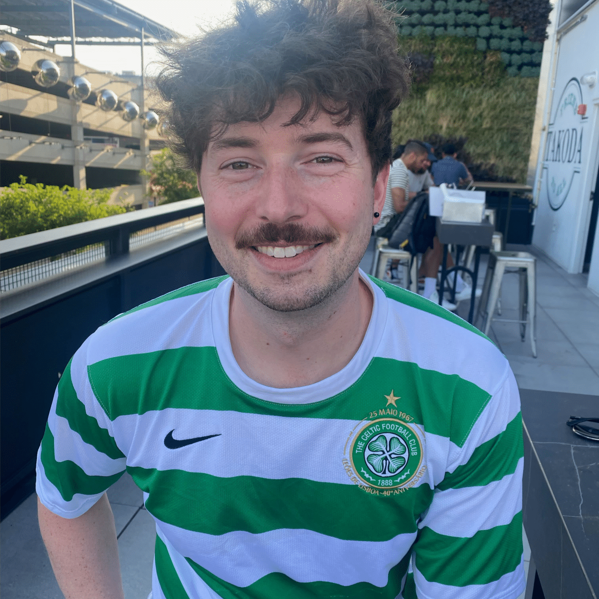 A white man with brown curly hair and a mustache wearing a white and green striped Nike shirt and he is smiling.