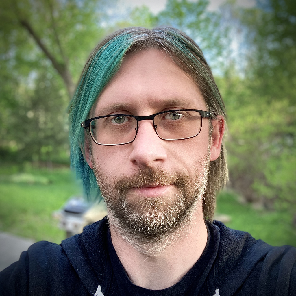 A photograph of Jeremy Wagner. He is standing against a backdrop of grass and trees, with a green-to-blue swash on the left side of his hair.