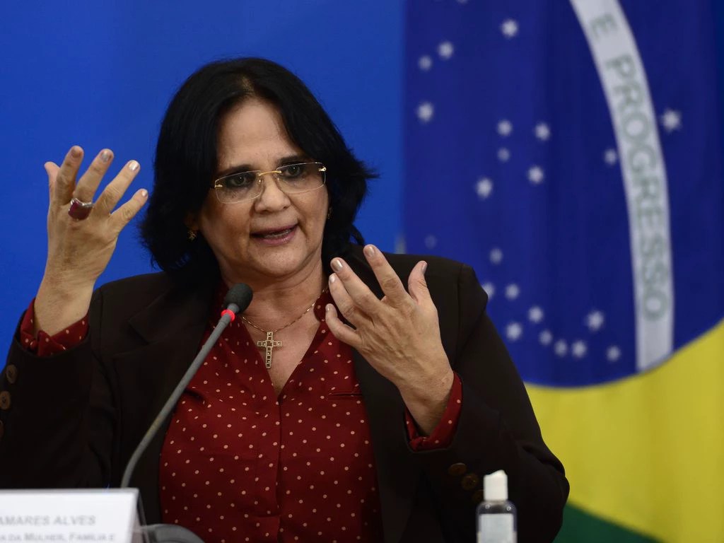 Ex ministra da Mulher durante o governo Bolsonaro e hoje senadora, Damares Alves. A política é uma mulher branca de olhos e cabelos escuros. Ela usa óculos de grau e veste blazer marrom com camisa social vermelha de poá. Ela usa uma corrente dourada com um pingente de cruz