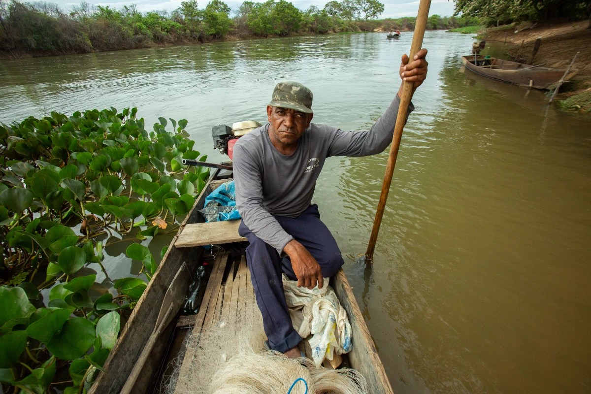 Homem negro posa sentado em um barco à beira de um rio; ele veste calça e blusa de mangra longa cinza, um boné verde escuro e segura na mão direita um bastão de madeira