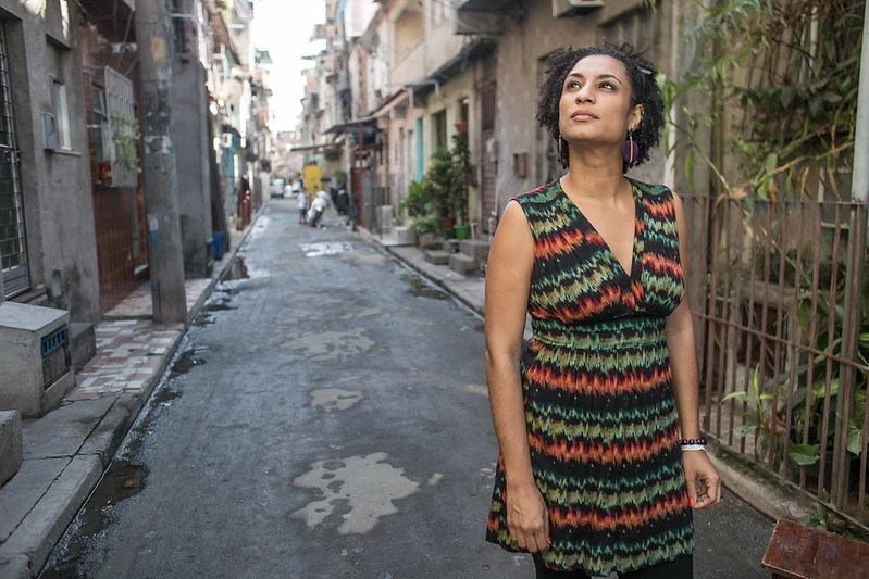 Marielle Franco era uma mulher negra com cabelos castanhos; na imagem ela está parada em uma rua da favela da Maré, usando um vestido colorido