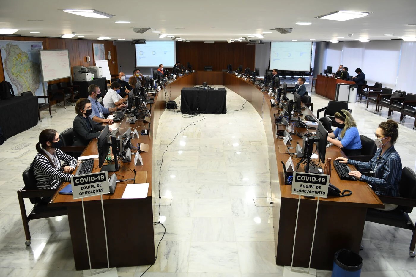 Equipe de planejamento do governo federal no combate ao coronavírus está sentada em duas fileiras de mesas paralelas, trabalhando no computador