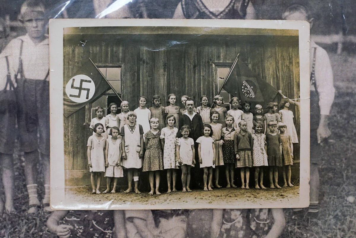 Imagem exposta no corredor da secretaria de Educação de Dona Emma, na imagem, crianças com um adulto posam para a foto em frente às bandeiras do Brasil e uma bandeira estampada com uma suástica nazista.