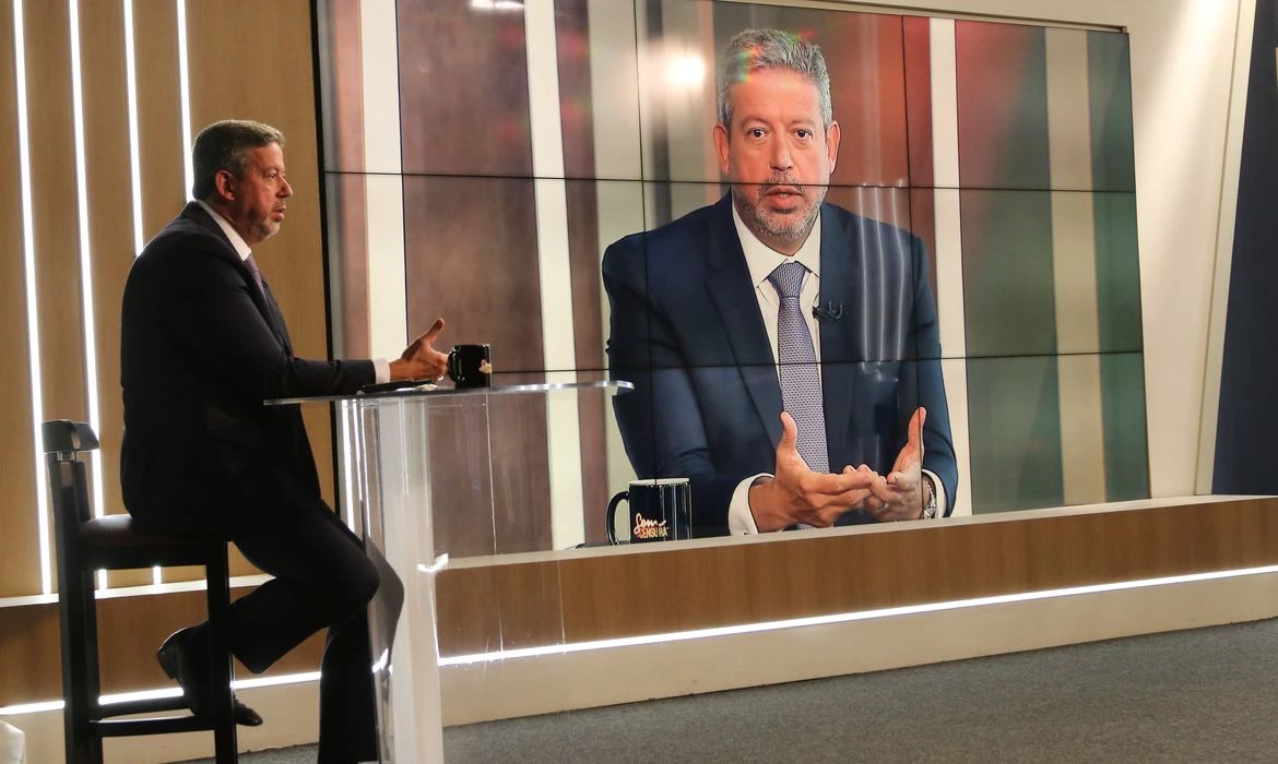 Presidente da Câmara, Arthur Lira, é um homem branco com cabelos e barbas grisalhos. Na imagem ele está sentado de lado e sua imagem é refletida em um telão atrás dele. Lira fala ao microfone e gesticula