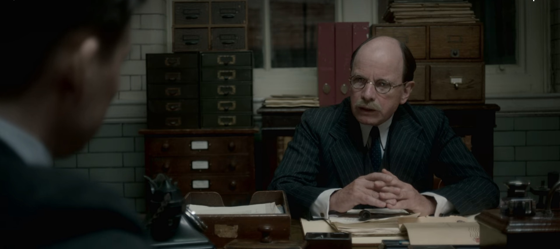Screen grab from The Crown showing an actor playing a bald and bespectacled Atlee at his desk in his office. Behind him on a credenza are a variety of files including two card index boxes each with four drawers.