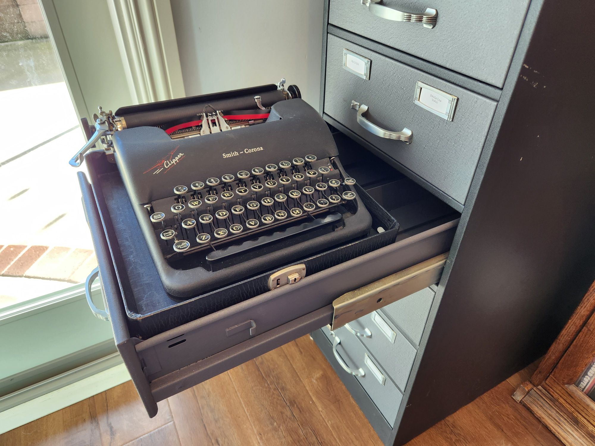 A drawer is pulled out of the filing cabinet and a black typewriter sits on top of it as an impromptu typing desk.