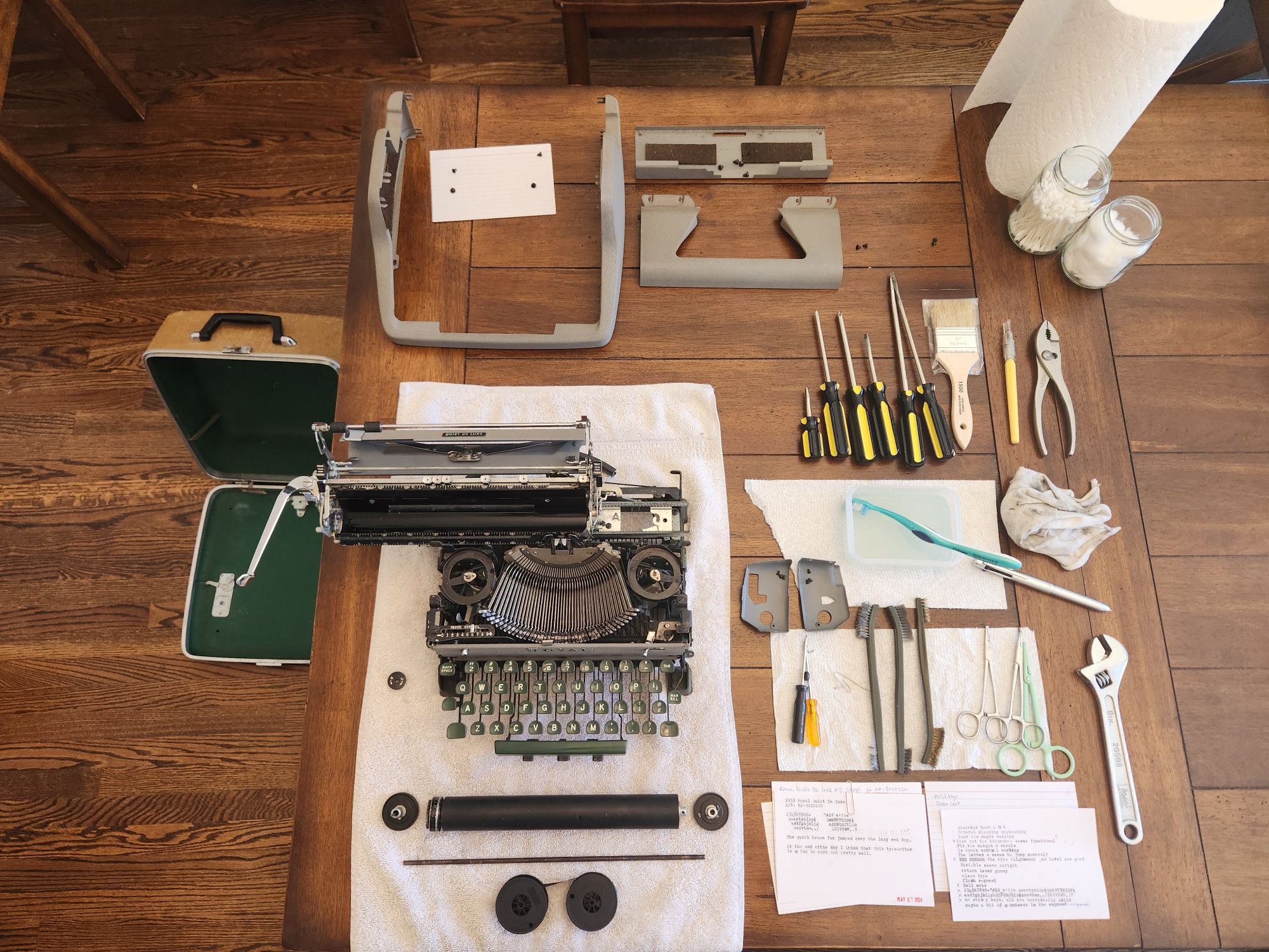 A table laid out with a white towel on which sits the dismantled skeleton of a Royal typewriter. Around it are ribbon spools, a platen and knobs, the various parts of the exterior body, tools, brushes, and repair instruments.
