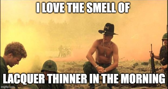 Screencapture from the film Apocalypse Now featuring a shirtless man with a calvary-style military hat squatting down  on a beach while two soldiers work in front of him. He is literally surrounded by the yellow fog of war. The meme image is surrounded with the words: I love the smell of lacquer thinner in the morning.