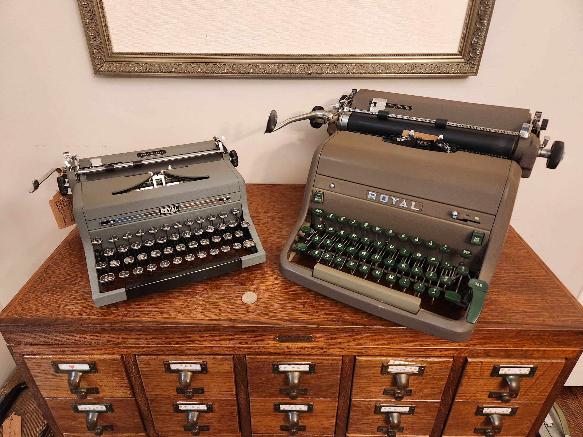 A straight on view of a small gray Royal QDL typewriter next to a brown Royal HH which is almost 2 1/2 times the size of  the first. Both sit on top of a wooden library card catalog.