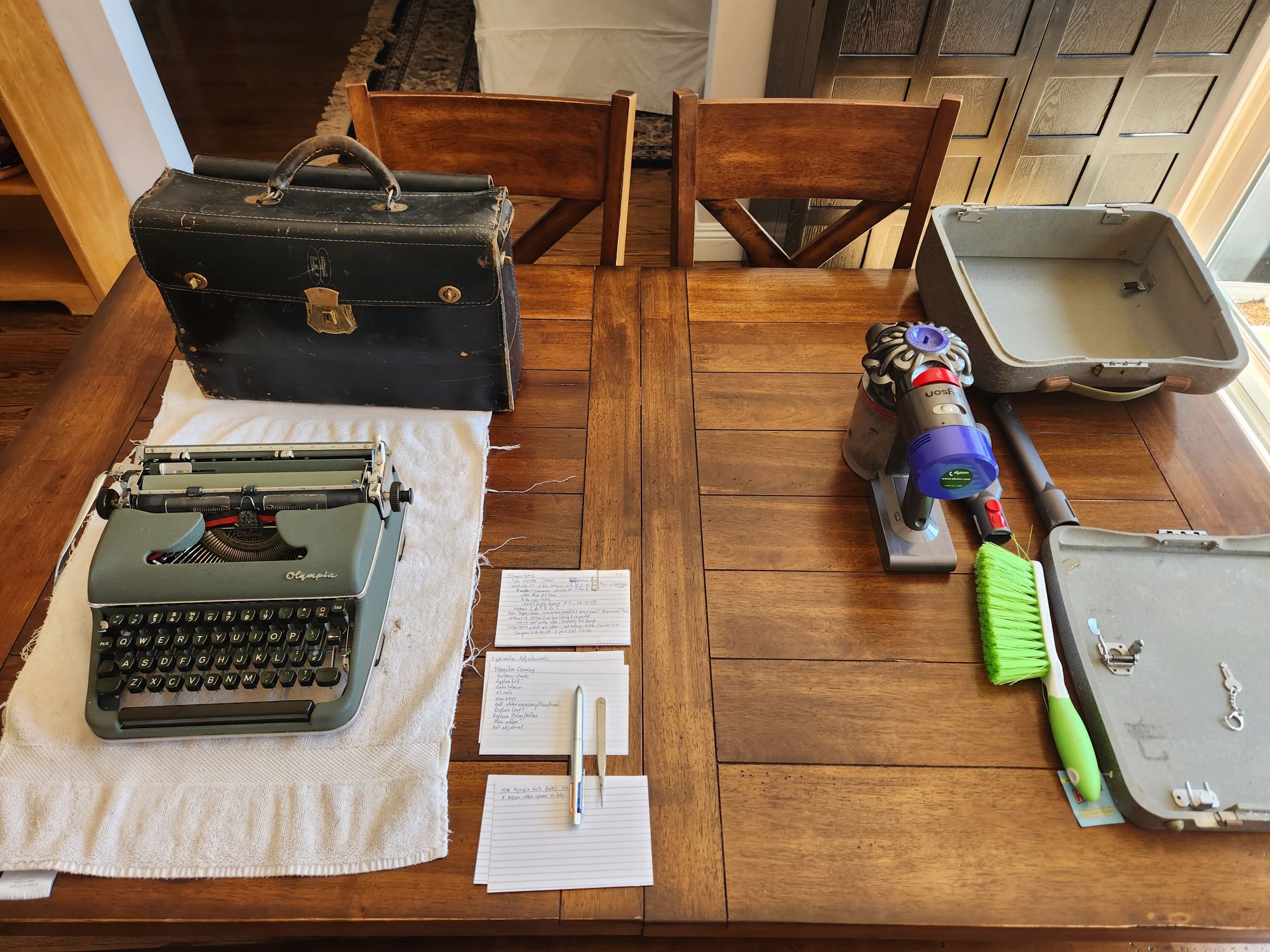 A black leather tool case has appeared on the table above the typewriter which now sits on the hand towel next to some index cards and a pen. The case and vacuum/brush have been pushed off to the right side of the table.