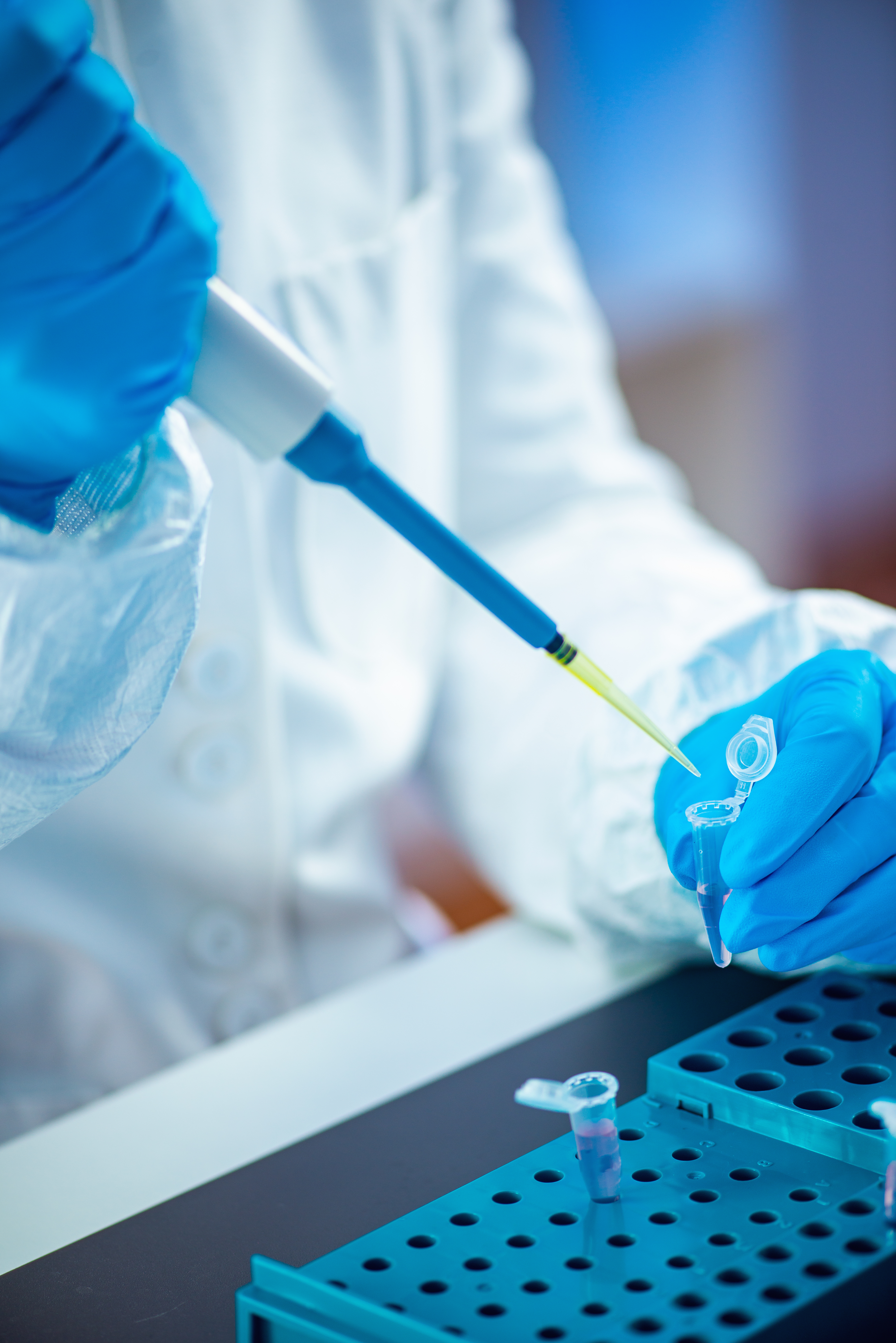 The image shows a scientist in a lab coat and gloves holding a pipette filled with a clear liquid. The pipette is inserted into a test tube filled with a yellow liquid. The scientist is wearing a lab coat and gloves, and is holding a pipette filled with a clear liquid. The pipette is inserted into a test tube filled with a yellow liquid.