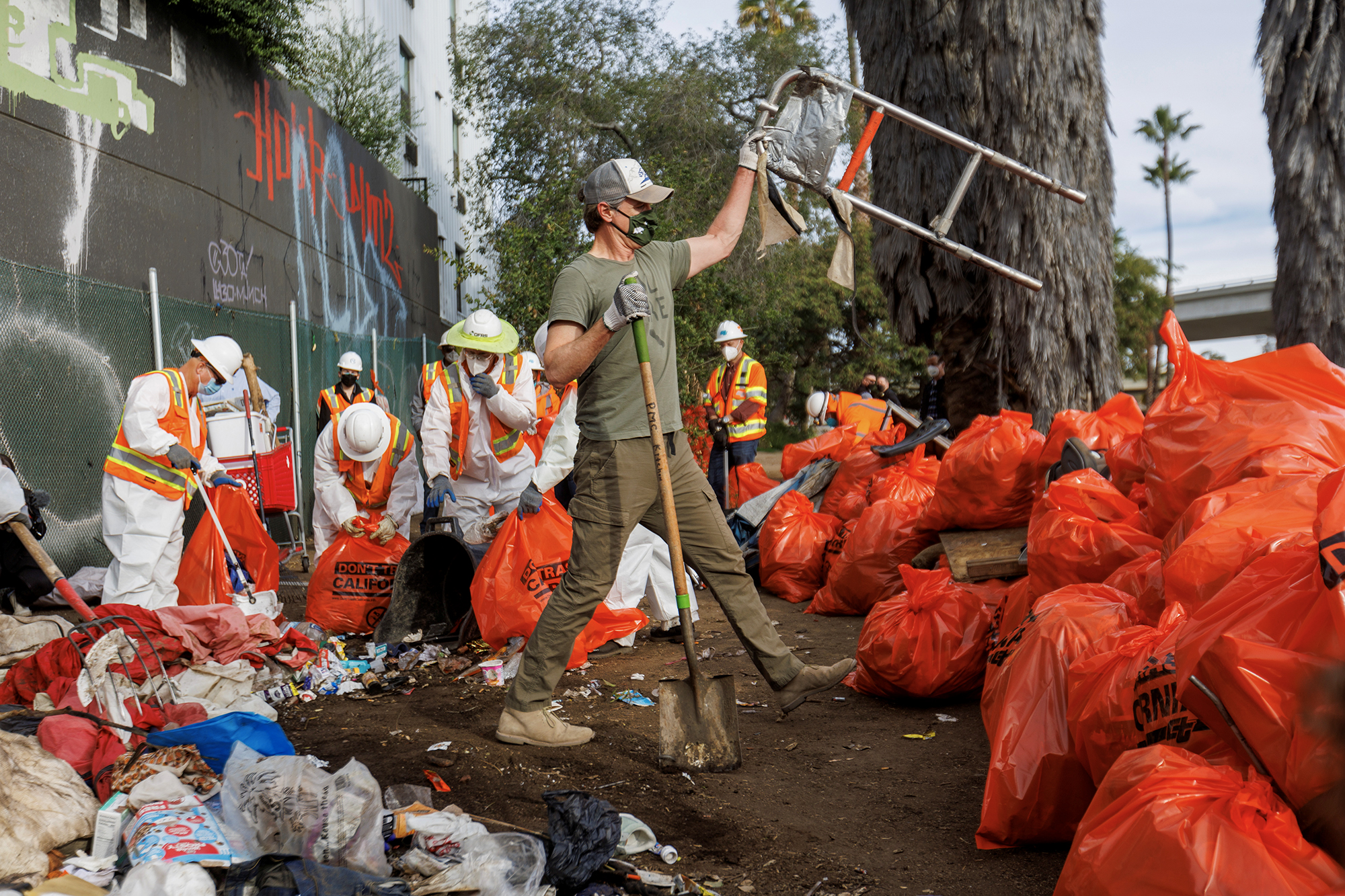 Surrounded by workers in hazmat suits and bright orange waste bags, Gov. Gavin Newsom helps clear a homeless encampment in San Diego.