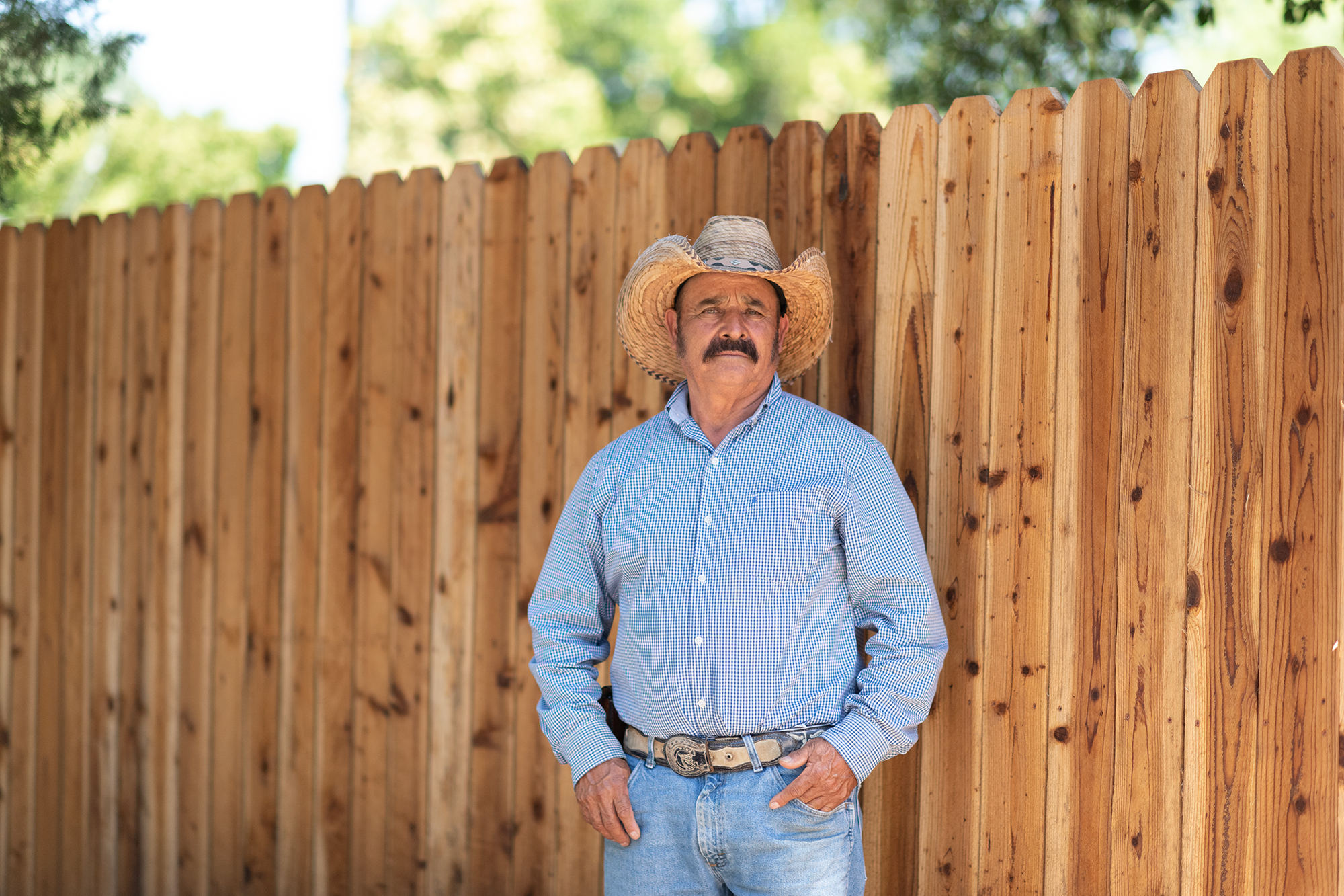 Farmworker Isidro Fierros Hernandez, wearing a cowboy hat, blue plaid shirt and blue jean, stands near a fence at City Park in Winters on June 10, 2024. Photo by Laure Andrillon for CalMatters