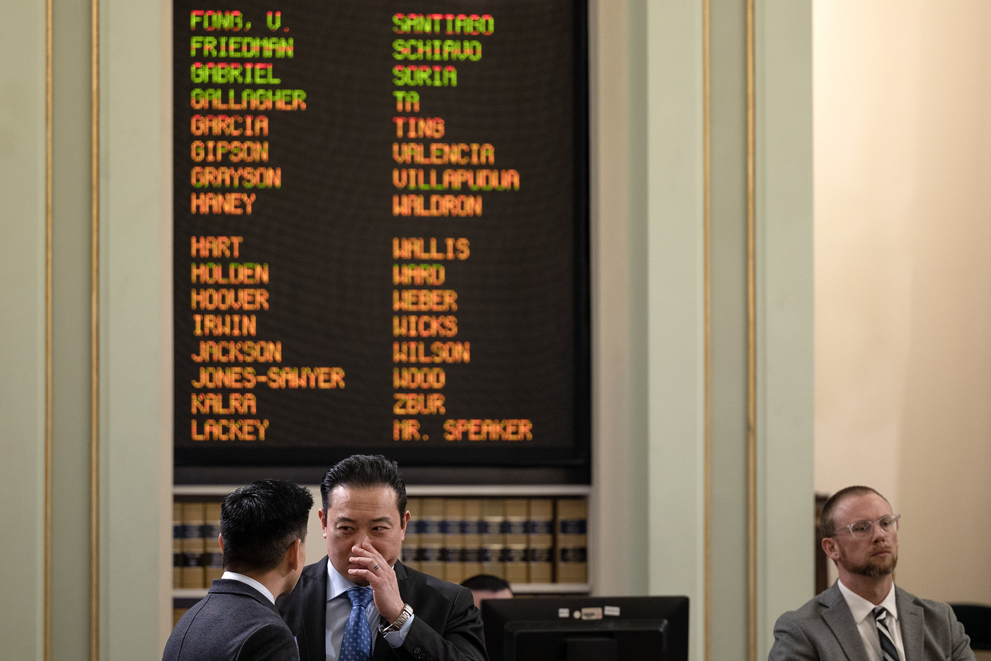 Two individuals are engaged in a private conversation in the foreground of a legislative chamber, with one appearing to whisper to the other. A digital display board in the background lists names in green, red, and orange, indicating legislative votes or attendance. A third person in a suit and glasses sits nearby, observing the scene. Bookshelves and muted green walls with gold accents frame the setting.