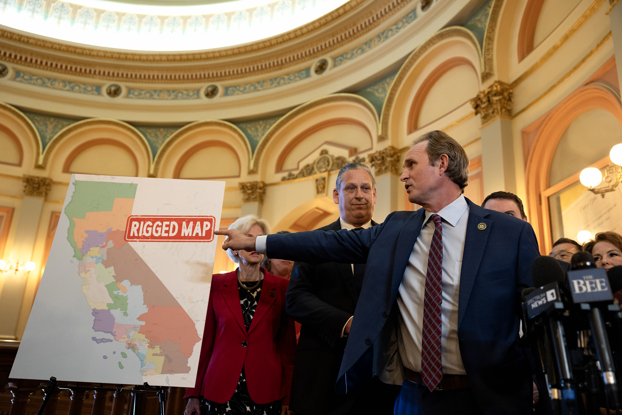 A person in a suit gestures toward a large poster of California’s district map labeled “Rigged Map” in red text, during a press conference inside a government building with ornate arches and a domed ceiling. Several people and news microphones are visible around them.