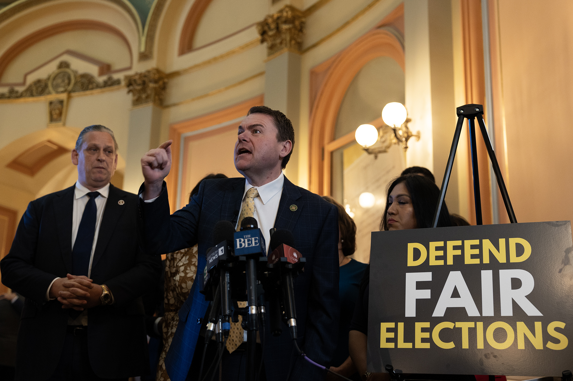 A person in a suit speaks emphatically at a podium surrounded by microphones, with others standing nearby. A large sign next to the podium reads, "Defend Fair Elections."